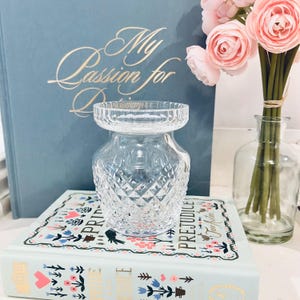 May include: A clear, diamond-cut glass vase with a wide top and a rounded body. The vase sits atop a book with floral designs and the title "Pride and Prejudice." A book with the title "My Passion for" is in the background, along with pink flowers in a glass vase.