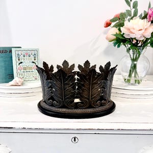 May include: A dark bronze metal crown-shaped decorative piece with detailed leaf design. The crown sits on a circular base. It is displayed on a white surface. A vase of flowers and books are in the background.