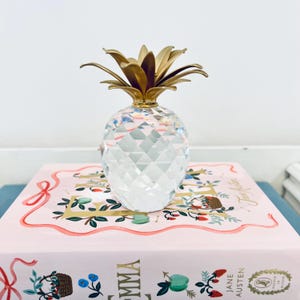 May include: A clear crystal pineapple ornament with a gold-coloured crown sits on a pink book with floral illustrations and the title "Emma". The pineapple has a faceted design, reflecting light.