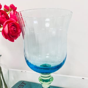 May include: A tall, clear glass with a light blue bowl and a turquoise base. The stem is a light green color. The rim of the glass has a wavy design. The glass is next to a bouquet of red flowers.