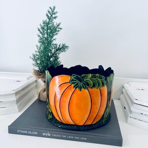 May include: A ceramic pumpkin-shaped container with an orange exterior and green leaves. The container sits on a book with the text "style create the look you love". A small evergreen tree is in the background.
