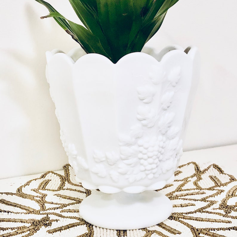 May include: White milk glass planter with a scalloped rim and embossed grape design. The planter has a pedestal base and is filled with a green plant.