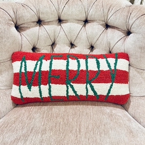 May include: A rectangular decorative pillow with red and white stripes. The word "MERRY" is embroidered in green across the center. The pillow is placed on a tufted beige armchair, suggesting a holiday theme.