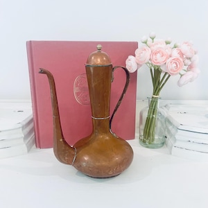 May include: A tall copper pitcher with a long, slender spout, a curved handle, and a rounded base. The pitcher features a removable lid with a small knob at the top. The metal surface shows a warm, metallic brown patina with subtle variations.
