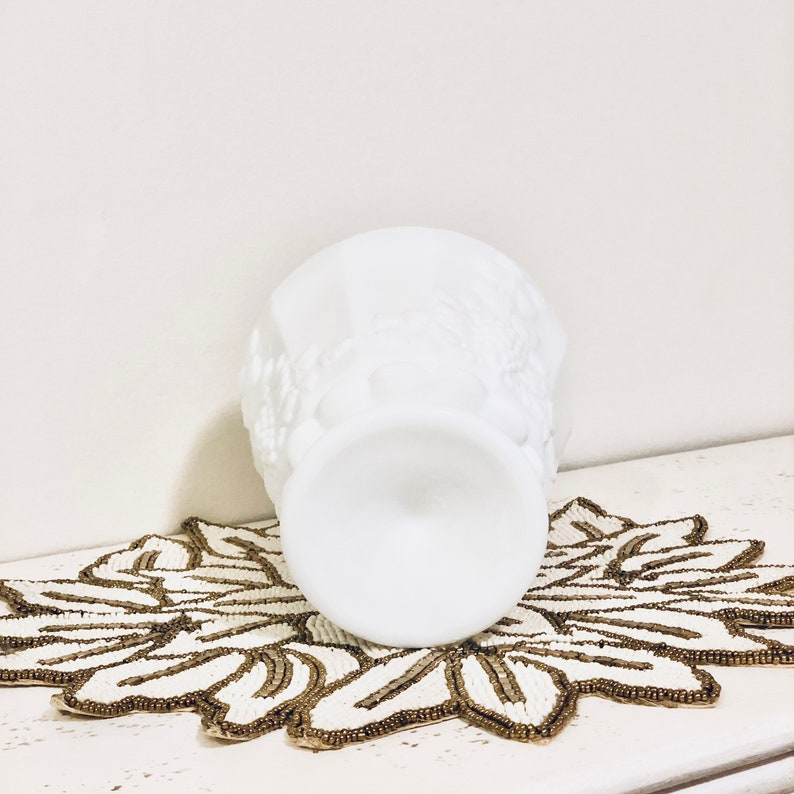 May include: White milk glass vase with a floral design. The vase is sitting on a beaded doily.