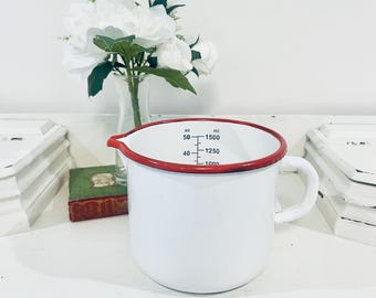 May include: A white enamel measuring pitcher with a red rim and a side handle. The interior features black measurement markings in ounces and milliliters, ranging from 40 to 50 ounces and 1000 to 1500 milliliters, with a pouring spout on the rim.