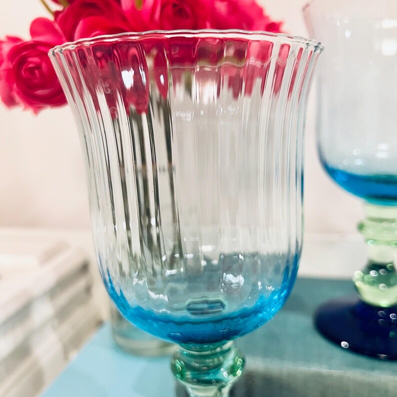May include: A clear glass goblet with a blue base and a green stem. The glass has vertical ridges and is partially filled with water. Pink flowers are visible in the background, adding a pop of color.