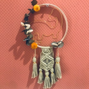 Peut inclure: Une suspension murale en macramé avec un cercle en bois décoré de feuilles d'eucalyptus séchées, de petites fleurs blanches et de pompons jaunes. Le macramé présente un motif géométrique avec des glands.