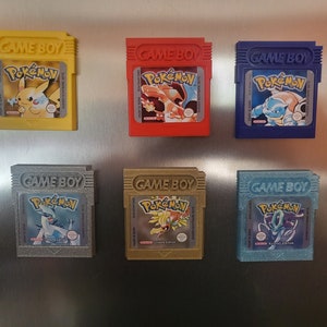 Könnte beinhalten: Sechs verschiedene farbige Game Boy-Module für das Videospiel Pokémon. Die Module sind in einer Reihe auf einer silbernen Oberfläche angeordnet. Die Module sind alle in verschiedenen Farben, darunter Gelb, Rot, Blau, Grau, Gold und Hellblau.