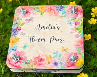 May include: A wooden flower press with the text "Amelia's Flower Press" in black script. The press is decorated with a colorful floral border featuring pink, yellow, and blue flowers, and butterflies. Four metal bolts secure the press.