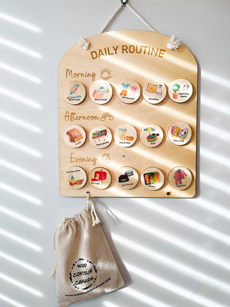 May include: A wooden daily routine chart with a rounded top and a hook for hanging. The chart is divided into three sections: Morning, Afternoon, and Evening. Each section has a sun, rainbow, and moon icon respectively. The chart has 15 round wooden pieces with images and text representing daily activities like brushing teeth, getting dressed, lunch, playing, and bedtime. The chart is made by Wood Contour Canada.