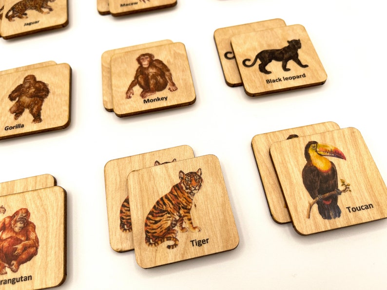 Op de afbeelding: Houten vierkante tegels met illustraties van dieren, waaronder een gorilla, een aap, een tijger en een toekan. Elke tegel heeft de naam van het dier eronder afgedrukt. De tegels zijn gemaakt van lichtgekleurd hout.