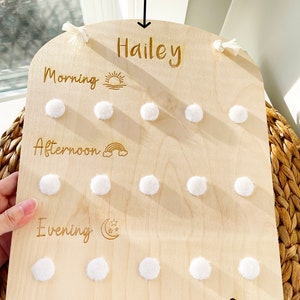 May include: A wooden board with a rounded top and a white string attached to each side. The board has a white background and is engraved with the words 'Personalization Option', 'Hailey', 'Morning', 'Afternoon', 'Evening', and a sun, rainbow, and moon with stars. There are white pom poms attached to the board in three rows of six.