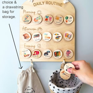 May include: A wooden daily routine board with 28 round wooden tokens. The board is light brown and has the words 'DAILY ROUTINE' written in gold lettering. The board is divided into three sections: Morning, Afternoon, and Evening. Each section has a different icon and the words 'Brush teeth', 'Wash hands', 'Get dressed', 'Breakfast', 'Watering Plants', 'Lunch', 'Plant garden', 'Snack time', 'Pool', 'Story time', 'Put toys away', 'Get mail', 'Care for pet', 'Dinner', and 'Pajamas' written on the tokens. The board has two hooks on the bottom. A small beige drawstring bag with the words 'WOOD CONTOUR CANADA' and a website address is hanging on one hook. A small beige and white basket with a black and white cross pattern is hanging on the other hook. Two tokens are being held in a hand. The tokens have the words 'Take out trash' and 'Put toys away' written on them.
