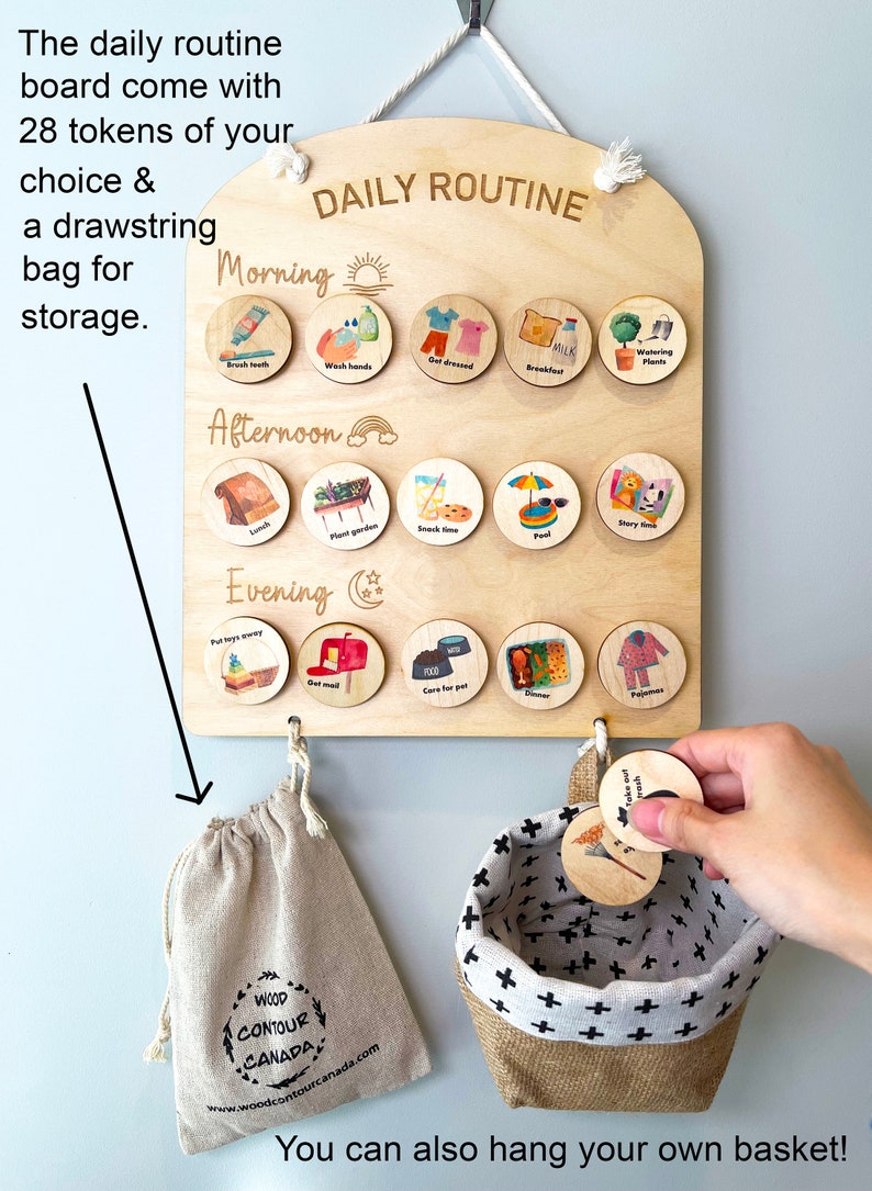 May include: A wooden daily routine board with 28 round wooden tokens. The board is light brown and has the words 'DAILY ROUTINE' written in gold lettering. The board is divided into three sections: Morning, Afternoon, and Evening. Each section has a different icon and the words 'Brush teeth', 'Wash hands', 'Get dressed', 'Breakfast', 'Watering Plants', 'Lunch', 'Plant garden', 'Snack time', 'Pool', 'Story time', 'Put toys away', 'Get mail', 'Care for pet', 'Dinner', and 'Pajamas' written on the tokens. The board has two hooks on the bottom. A small beige drawstring bag with the words 'WOOD CONTOUR CANADA' and a website address is hanging on one hook. A small beige and white basket with a black and white cross pattern is hanging on the other hook. Two tokens are being held in a hand. The tokens have the words 'Take out trash' and 'Put toys away' written on them.