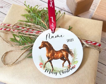 May include: A round Christmas ornament featuring a brown horse illustration with the text "Merry Christmas" and "2023". The ornament is on a gift wrapped in brown paper, tied with twine and greenery.