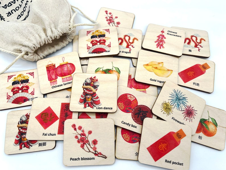 May include: A collection of square wooden tiles featuring colorful illustrations of Lunar New Year symbols, including a lion dance, red pockets, gold ingots, Chinese lanterns, and peach blossoms, with English and Chinese text labels on each tile.