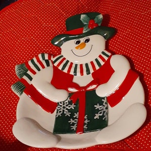 May include: A ceramic snowman-shaped serving plate with a green hat, red vest, and striped scarf. The snowman holds a green gift box decorated with snowflakes and a red bow. The plate is white with red and green accents, perfect for holiday gatherings.