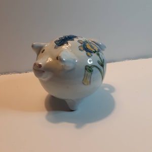 May include: A white ceramic piggy bank with blue, yellow, and green floral designs painted on the side.