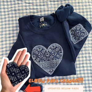May include: A navy blue crewneck sweatshirt featuring a large white heart-shaped graphic with intricate patterns on the chest and matching heart-shaped elbow patches on the sleeves. The text reads ELBOW PADS CHANGE and UPDATED BELOW PADS ARE THESE HEART ONES.