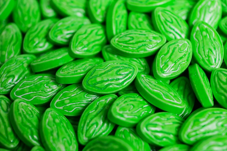 Puede incluir: Primer plano de un mont&oacute;n de caramelos verdes brillantes. Cada caramelo tiene forma ovalada con un dise&ntilde;o blanco y arremolinado en la superficie. Los caramelos son brillantes y parecen caramelos duros o pastillas.