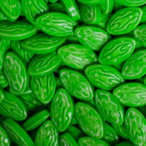Puede incluir: Primer plano de un mont&oacute;n de caramelos verde brillante. Cada caramelo tiene forma ovalada con una superficie ligeramente texturizada y un dise&ntilde;o blanco en espiral. Los caramelos est&aacute;n dispersos, llenando el marco y creando una exhibici&oacute;n vibrante y llamativa.
