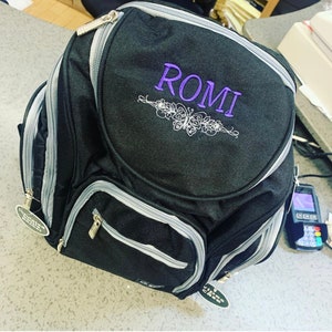 May include: A black diaper bag with silver accents and a zippered pocket. The bag has a purple embroidered design with the name "ROMI" on the front.