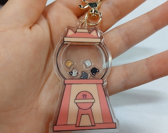Peut inclure: Un porte-clés en acrylique transparent en forme de distributeur de chewing-gum avec une base rose et un dessus transparent rempli de petites figurines de chats. Le porte-clés est doté d'un fermoir en métal doré.