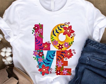 May include: White t-shirt with a colorful floral "LOVE" graphic. The shirt is folded and displayed with a pair of brown ankle boots and blue jeans.