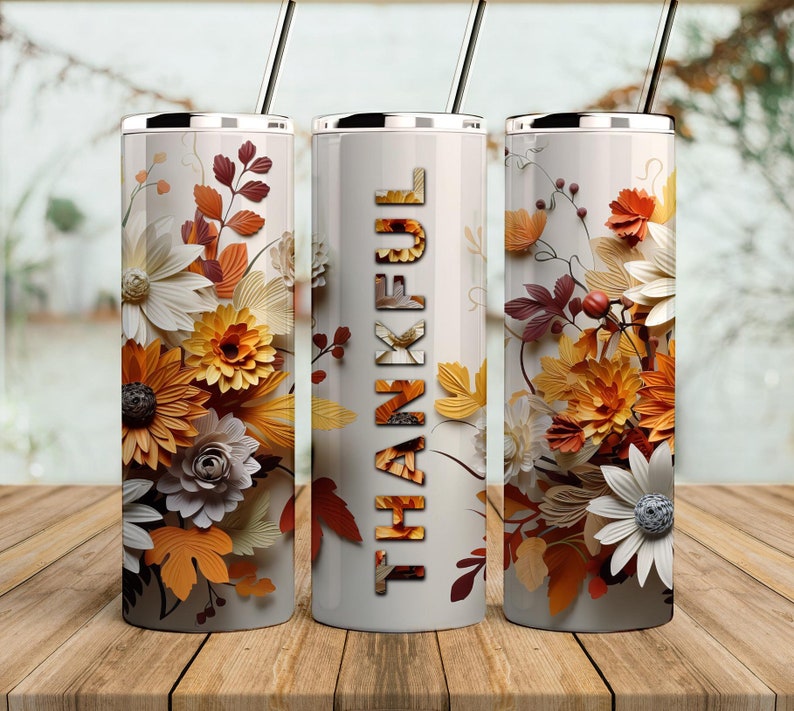 May include: Three stainless steel tumblers with lids and straws. The tumblers are decorated with a floral design featuring white, yellow, and orange flowers and leaves. The middle tumbler has the word "THANKFUL" printed in black letters.