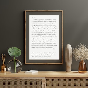 Puede incluir: Una impresi&oacute;n en blanco y negro enmarcada con una carta escrita a mano en letra cursiva. La carta est&aacute; sobre un fondo blanco y est&aacute; enmarcada en un marco de madera marr&oacute;n. La impresi&oacute;n est&aacute; sobre una mesa de madera con un jarr&oacute;n de flores blancas y un jarr&oacute;n de vidrio con una planta verde.
