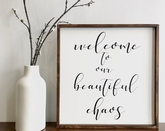 May include: A wooden sign with a white background and black calligraphy that reads "Welcome to our beautiful chaos". The sign is sitting on a wooden surface with a white vase and a branch with white flowers.