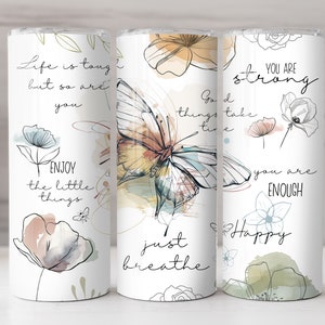 May include: Three white tumblers with inspirational quotes and watercolor floral designs. The quotes are "Life is tough but so are you", "Good things take time, just breathe", and "You are strong, you are enough, Happy".