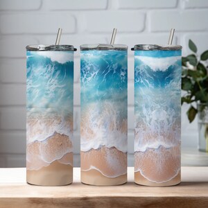 May include: Three stainless steel tumblers with a beach scene design. The tumblers feature a blue ocean with white waves crashing on a sandy beach.