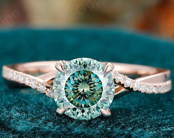 May include: A rose gold engagement ring with a large, round, teal gemstone. The band has a twisted design with small, clear accent stones. The ring is set against a teal velvet surface.