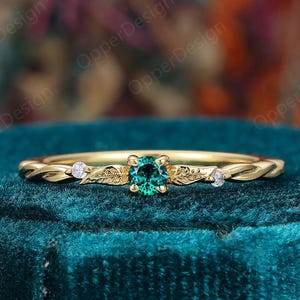 Op de afbeelding: Een gouden ring met een ronde groene middensteen geflankeerd door twee kleine heldere stenen. De ringband is ontworpen met gegraveerde bladmotieven aan weerszijden van de middensteen en een gedraaide metaalstructuur langs de rest van de ring.