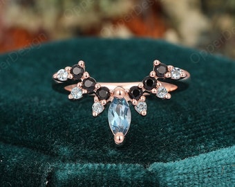 May include: A delicate rose gold ring with a teardrop-shaped aquamarine gemstone surrounded by smaller black and white diamonds. The ring is designed in a curved shape with a pointed top.