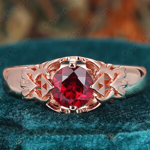 May include: Rose gold ring featuring a large, round, deep red gemstone. The ring has intricate side detailing and a polished finish. The gemstone is the focal point, set in a decorative setting. The ring is displayed on a dark green velvet surface.