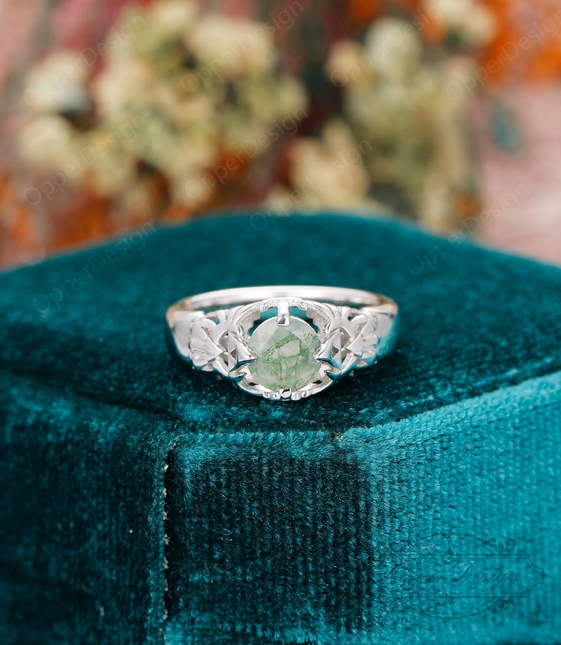 May include: A silver ring with a green gemstone set in a delicate, intricate design. The ring is on a teal velvet background.