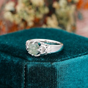 May include: A silver ring with a green gemstone set in a delicate, intricate design. The ring is displayed on a teal velvet box.