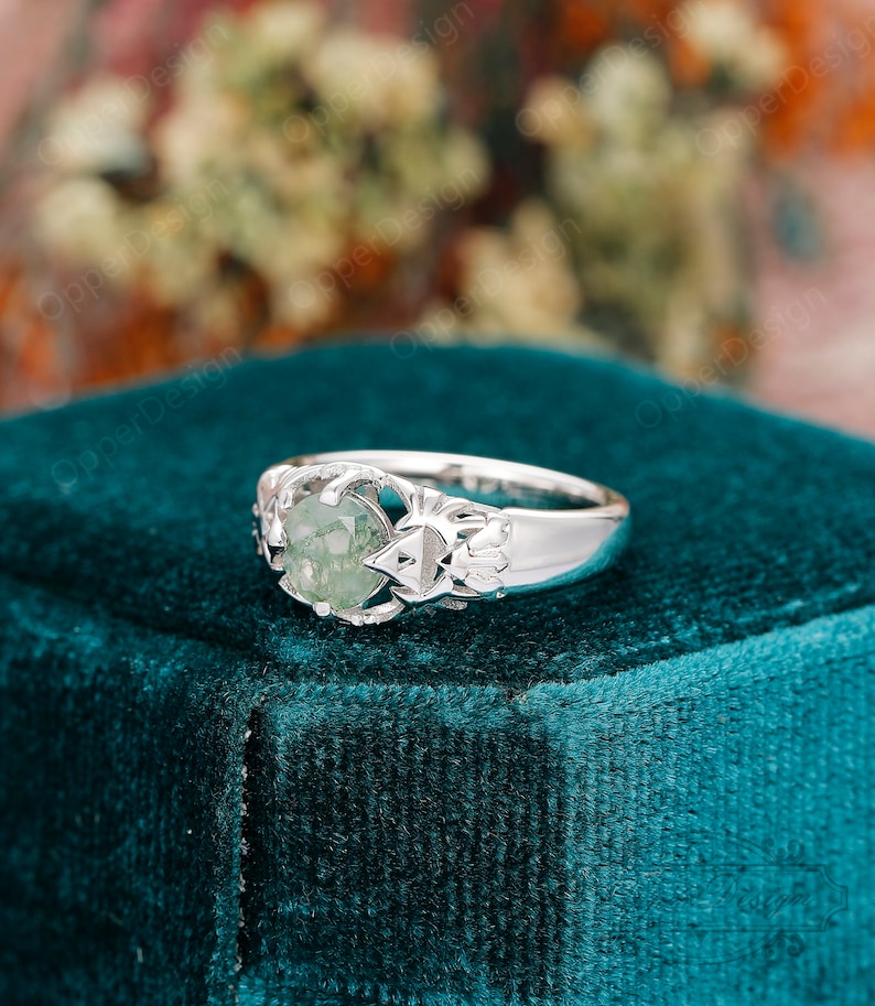 May include: A silver ring with a green gemstone set in a delicate, intricate design. The ring is displayed on a teal velvet box.