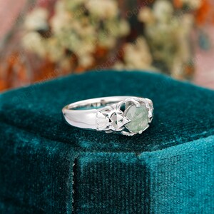 May include: A silver ring with a green gemstone set in a delicate, intricate design. The ring is on a teal velvet surface.