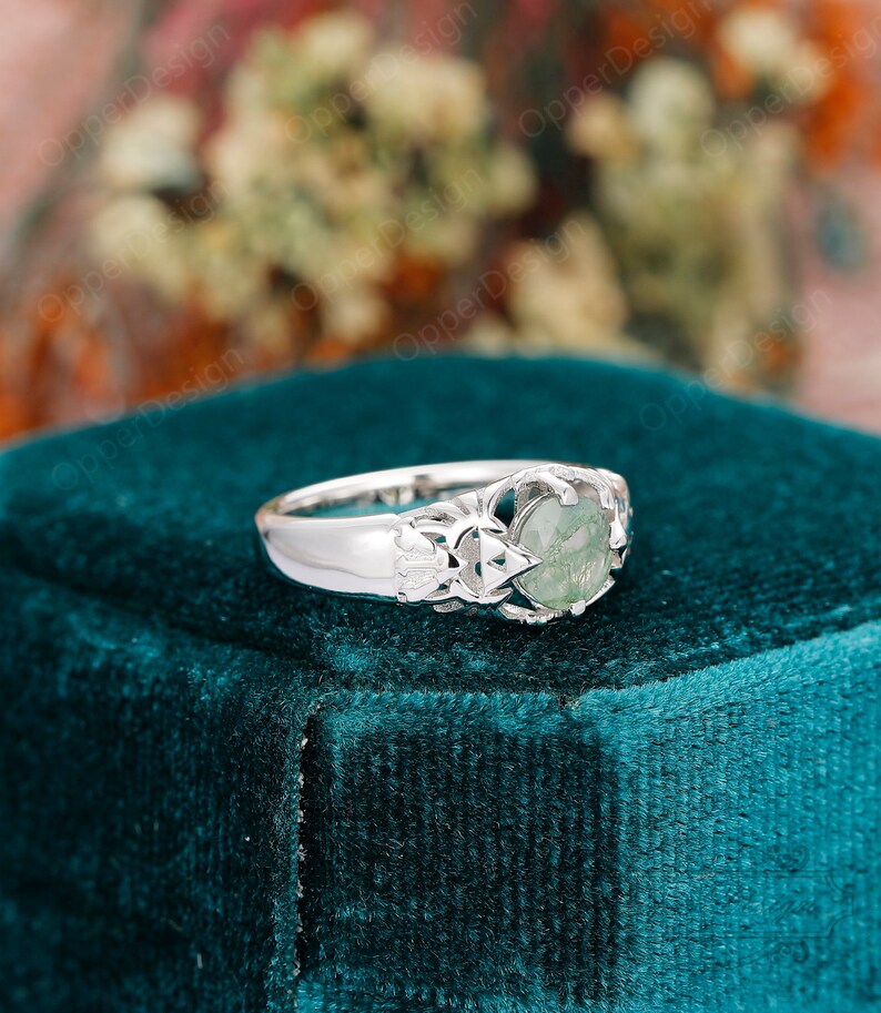 May include: A silver ring with a green gemstone set in a delicate, intricate design. The ring is on a teal velvet surface.