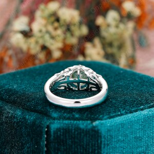 May include: A silver ring with a green gemstone set in a delicate, intricate design. The ring is on a teal velvet box.
