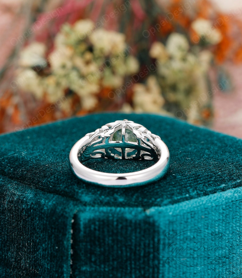 May include: A silver ring with a green gemstone set in a delicate, intricate design. The ring is on a teal velvet box.