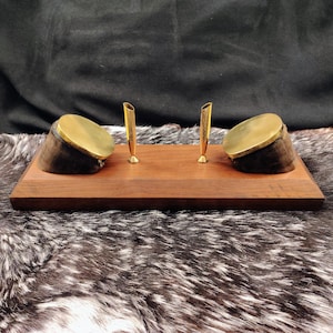 May include: A rectangular wooden desk stand featuring two dark, polished hoof-shaped accents topped with circular brass plates. Two slender, upright brass pen holders are positioned in the center of the smooth, dark brown wooden base.