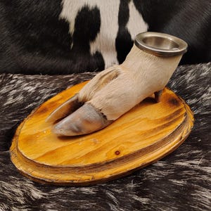 May include: A taxidermied animal foot with a metal ashtray on top, mounted on a wooden base. The foot is light brown and the base is a light wood color.