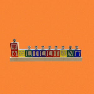 May include: A wooden Hanukkah menorah with the name "LILLIAN" spelled out in colorful blocks. The menorah features silver candle holders and traditional Jewish symbols. The background is a solid orange.