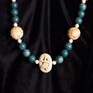 May include: A necklace featuring turquoise and cream-colored beads. The necklace has carved cream-colored beads interspersed with turquoise beads. The necklace is displayed on a black background.