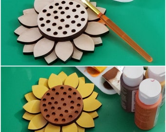 May include: A wooden sunflower craft with a brown center and a paintbrush. The sunflower has a circle of holes in the center. There are also paint bottles in the image.